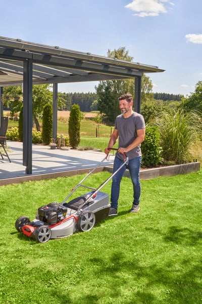 Ein Mann mäht an einem sonnigen Tag mit einem Benzinrasenmäher einen Garten vor einer modernen Terrassenüberdachung.