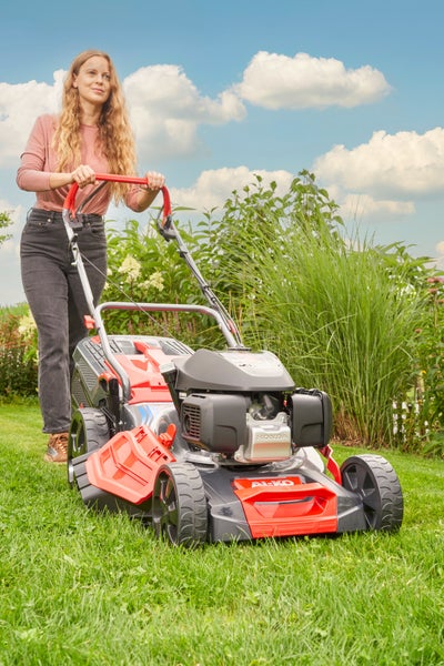 Eine Frau schiebt einen AL-KO Benzin Rasenmäher mit Honda Motor über eine Rasenfläche im Garten bei sonnigem Wetter.