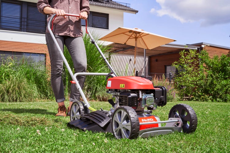 Eine Person nutzt einen AL-KO Benzin Rasenmäher zum Mähen auf einem Gartenrasen vor einem modernen Wohnhaus.