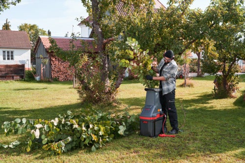 Ein Mann bedient einen elektrischen Gartenhäcksler auf einer Wiese im Garten und zerkleinert Äste.