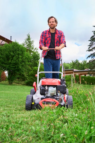 Ein Mann im karierten Hemd schiebt einen Benzinrasenmäher mit Verbrennungsmotor über eine Wiese in einem Garten.