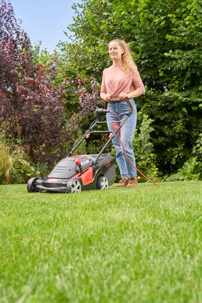Eine Frau nutzt einen elektrischen Rasenmäher der Marke AL-KO, um eine Rasenfläche vor einer Hecke im Garten zu mähen.