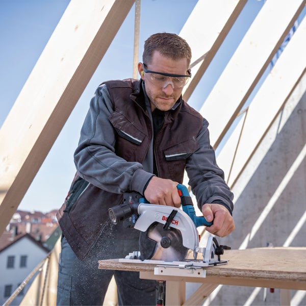 Handwerker mit Schutzbrille sägt eine Holzplatte mit einer Bosch Handkreissäge auf einer Baustelle unter einem Dachstuhl.