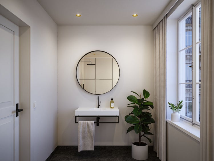 Modernes Badezimmer mit rundem Spiegel, wandhängendem Waschbecken mit schwarzem Metallgestell, schwarzer Armatur und grüner Zimmerpflanze.