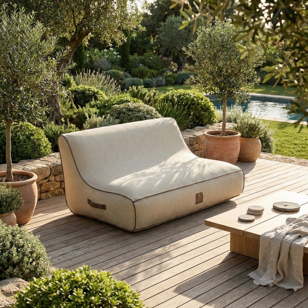 Helles Zweisitzer Outdoor-Sitzsack-Sofa auf einer Holzterrasse in einem mediterranen Garten mit Pool, Olivenbäumen und Natursteinmauer.
