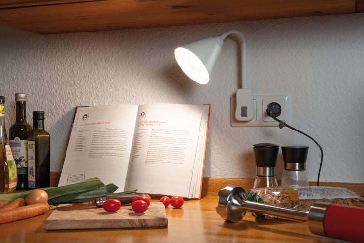 Küchenszene mit Kochbuch, Lebensmitteln, Schneidebrett, Stabmixer, Gewürzmühlen und einer weißen Arbeitsplatzleuchte