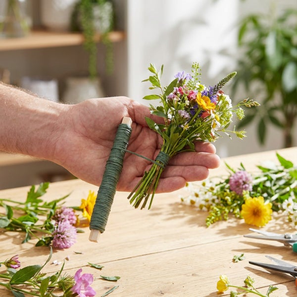 Hand hält einen kleinen Wildblumenstrauß, der mit Bindedraht auf einem Holzstab zusammengebunden wird, auf einem Holztisch mit weiteren Blumen.