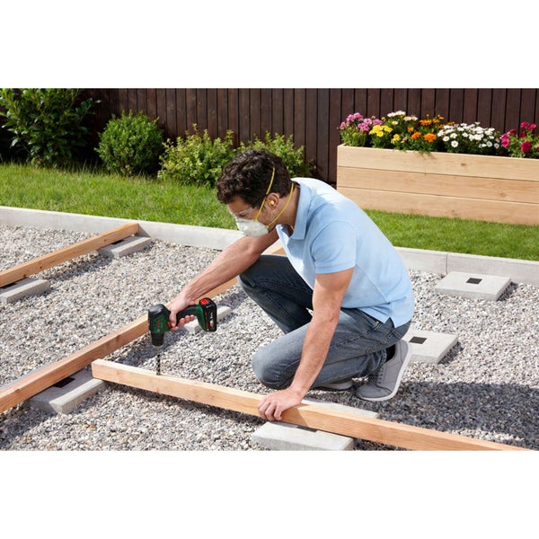 Ein Handwerker bohrt mit einem Bosch Akku Bohrhammer ein Loch in eine Holzunterkonstruktion auf einer mit Kies belegten Terrasse im Garten.