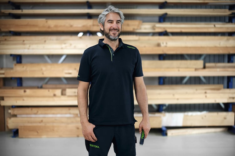Ein Handwerker mit grauen Haaren trägt ein Polohemd und Arbeitshose von Festool vor einem Holzlager im Hintergrund.