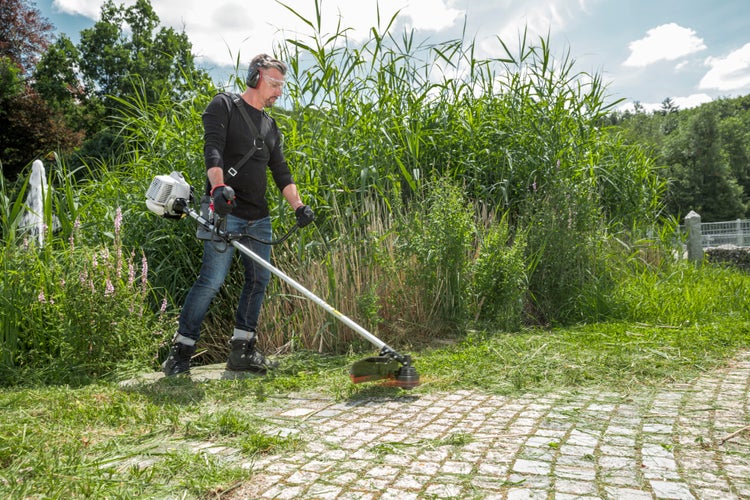 Ein Mann mit Schutzausrüstung nutzt einen Benzin Freischneider, um Gras an einem gepflasterten Weg neben einem Teich zu mähen.