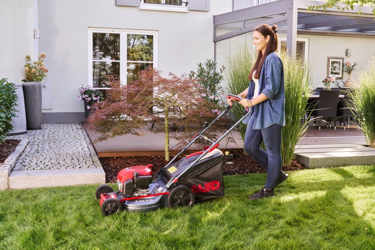 Eine Frau mäht den Rasen mit einem AL-KO Benzin-Rasenmäher in einem hellen, gepflegten Garten vor einem Wohnhaus.