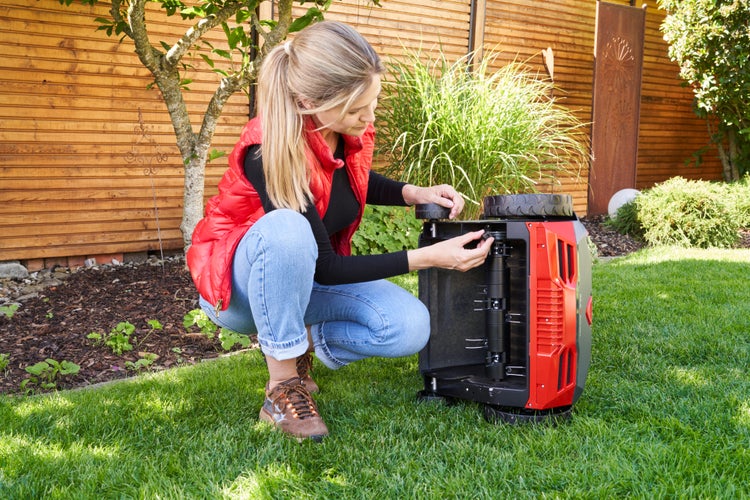 Eine Frau kniet im Garten auf dem Rasen und wartet die Unterseite eines Mähroboters.