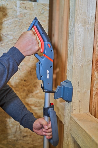 Eine Person hängt den Griff eines AL-KO Gartengeräts in eine Wandhalterung an einer Holzwand.