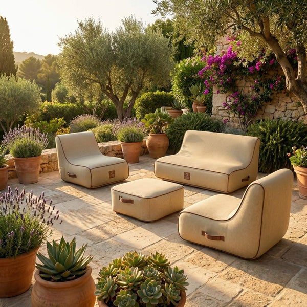 Garten-Lounge-Set aus Sesseln, Sofa und Hocker auf einer Steinterrasse in einem hellen, mediterran gestalteten Garten.
