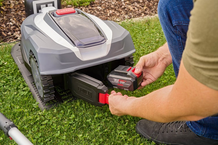 Person setzt einen 18 Volt oder 36 Volt Akku in das rückseitige Fach eines Mähroboters auf einer Rasenfläche ein.