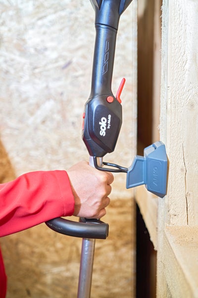 Eine Hand führt den Griff einer solo by AL-KO Motorsense in eine Wandhalterung an einer Holzwand ein.