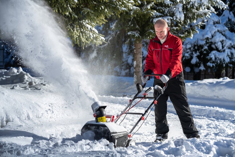 Eine Person bedient eine motorisierte Schneefräse, um eine verschneite Fläche vor Nadelbäumen zu räumen.
