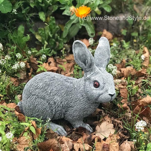Gartenfigur Hase aus Stein in sitzender Haltung platziert zwischen Laub und Pflanzen im Garten