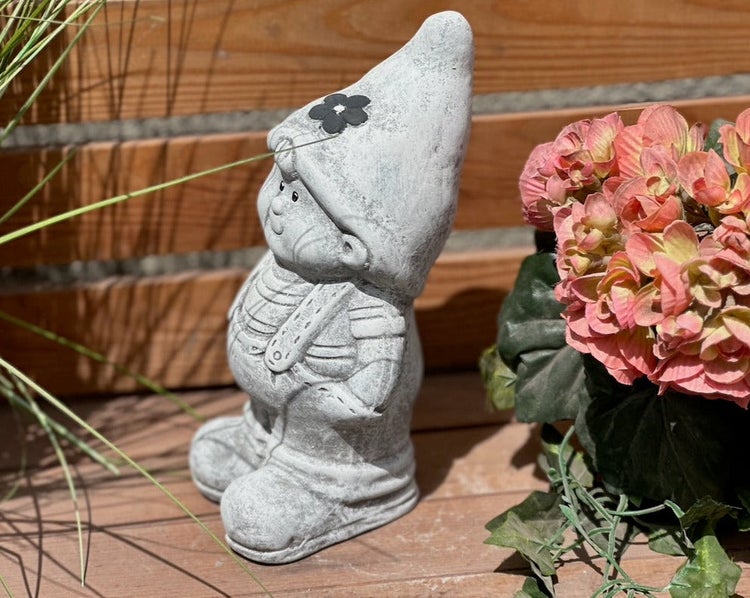 Gartenzwerg-Dekofigur aus grauem Stein mit Zipfelmütze und Latzhose, aufgestellt im Außenbereich neben einer Pflanze.