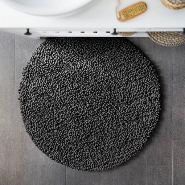 Runder Badeteppich in Anthrazit mit Chenille-Struktur vor einem weißen Waschtisch auf dunklen Fliesen.