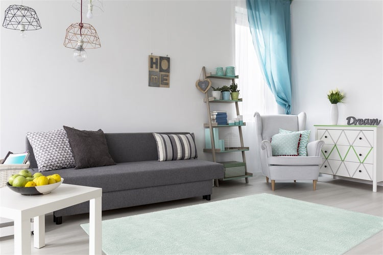Helles Wohnzimmer mit grauem Sofa, hellgrauem Sessel, mintgrünem Teppich, weißem Couchtisch und blauen Vorhängen in moderner Einrichtung.