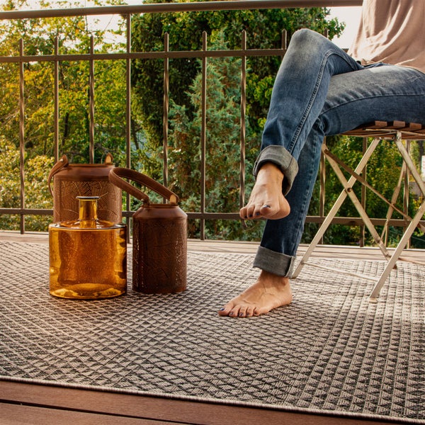 Außenteppich mit geometrischem Muster auf einem hellen Balkon mit Person auf Klappstuhl, Metalllaternen und einer Glasvase.