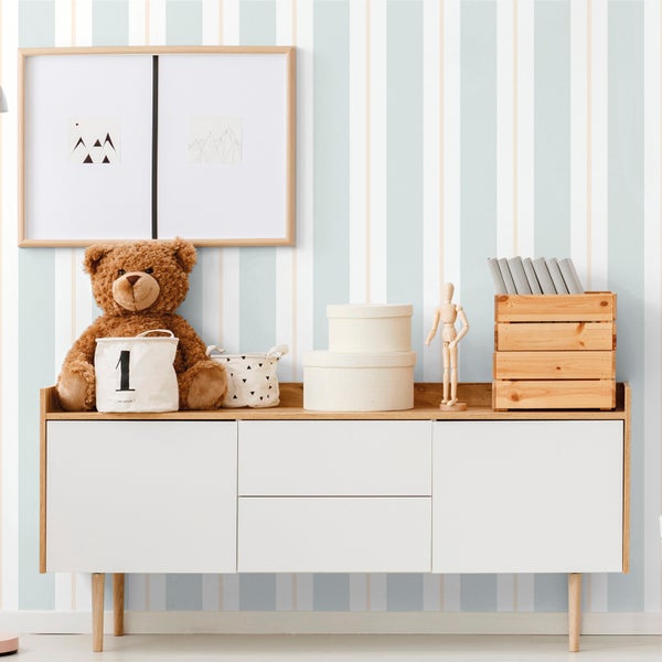 Helles Kinderzimmer mit vertikal gestreifter Vliestapete, einem Sideboard aus Holz mit weißen Fronten, einem Stoffbären und Aufbewahrungsboxen.