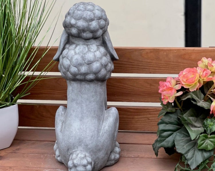 Gartenfigur Pudel aus Stein von hinten, sitzend auf einer Holzbank neben Pflanzen.