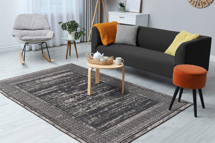 Moderner Wohnraum mit grauem Vintage-Teppich mit geometrischer Bordüre. Mit dunklem Sofa, Schaukelstuhl und orangefarbenem Hocker. Hell und ordentlich.