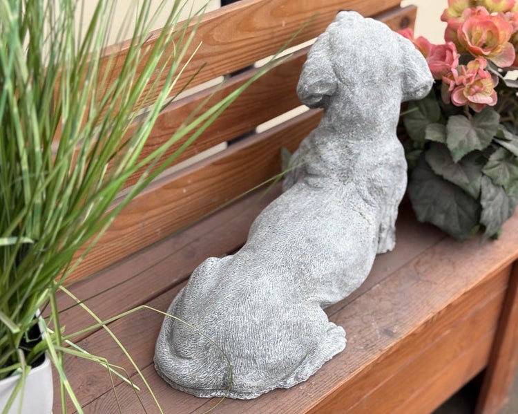 Gartenfigur liegender Hund aus Steinguss auf einer Holzbank neben Pflanzen.