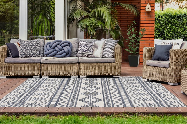 Möblierte Terrasse mit einem Rattansofa, Sesseln und einem geometrisch gemusterten Outdoor-Teppich auf einem Holzdeck mit Pflanzen.