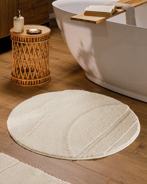 Runder cremefarbener Badteppich mit strukturiertem Linienmuster auf Holzboden neben freistehender Badewanne und Bambusbeistelltisch.