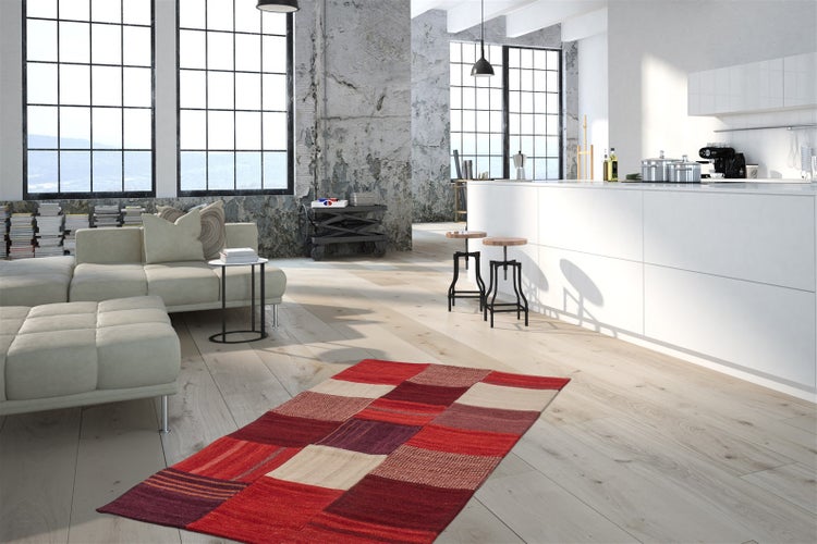 Moderner Industrie-Loft mit hellem Holzboden, rotem Patchwork-Teppich, weißer Kücheninsel und beigem Sofa. Große Fenster und Sichtbetonwände.