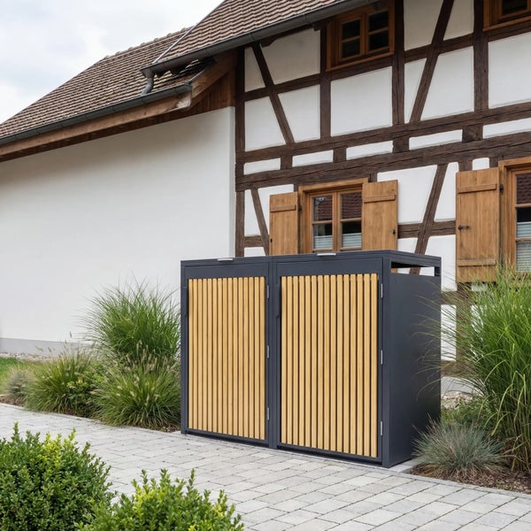 Moderne Mülltonnenbox aus Metall und Holzlamellen vor einem Fachwerkhaus auf gepflastertem Boden mit Ziergräsern.
