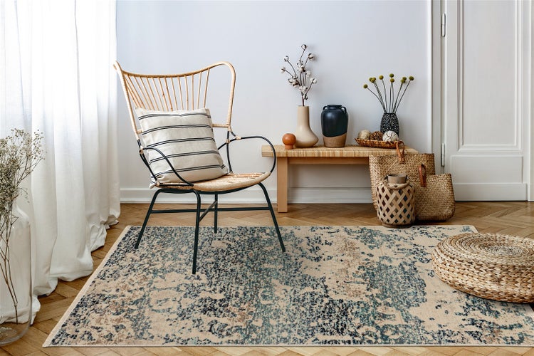 Heller Wohnraum mit einem gemusterten Teppich in Blau und Beige, einem Korbsessel mit Streifenkissen, einer Holzbank und geflochtenen Körben.