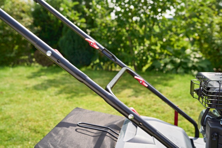 Detailansicht eines Rasenmäher-Führungsholms aus Metall mit roten Schnellverschlüssen in einem Garten.