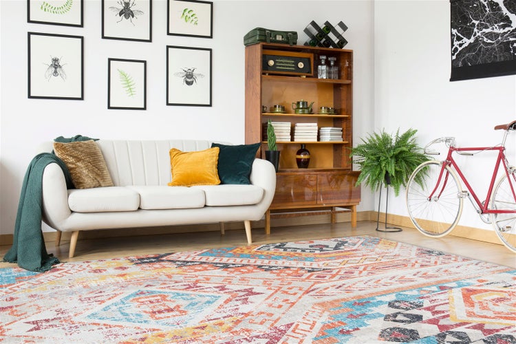 Wohnzimmer mit buntem Teppich mit geometrischem Muster, hellem Sofa, Holzregal und Wandbildern.