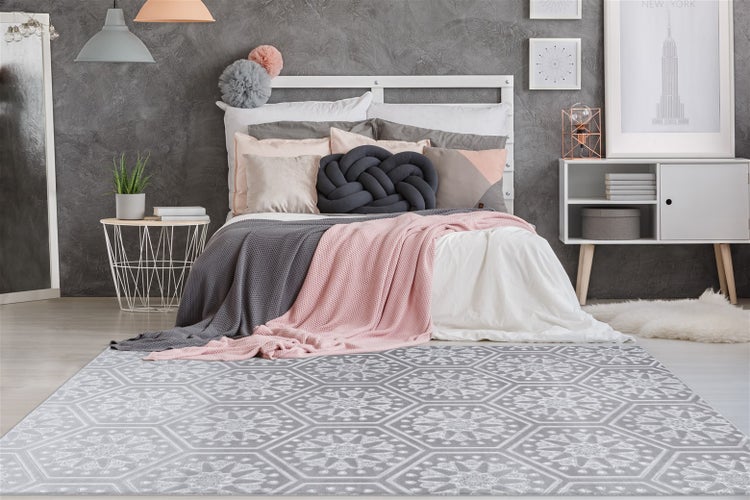 Modern eingerichtetes Schlafzimmer mit grau-weißem Musterteppich im Vordergrund, weißem Bettgestell und grauer Wand.