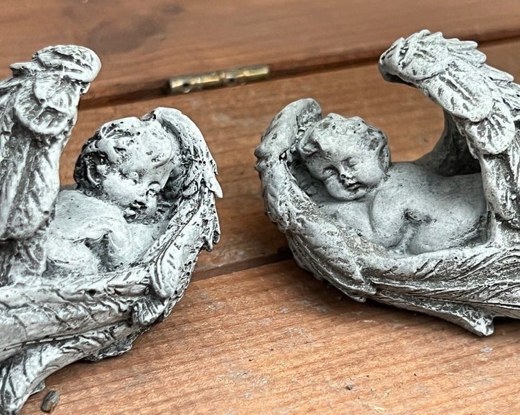 Zwei kleine Engelsfiguren aus Stein in liegender Position auf einer hölzernen Oberfläche.