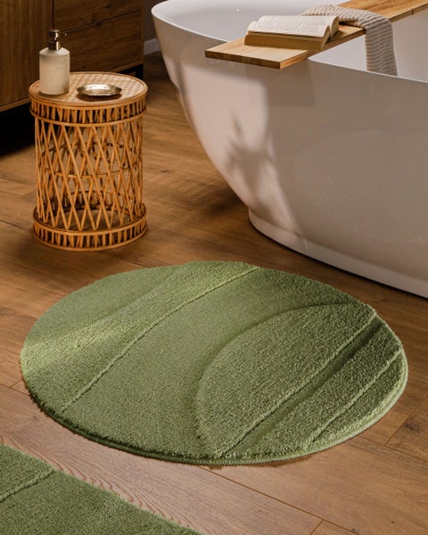 Runder grüner Badeteppich mit Wellenstruktur auf Holzboden in hellem Badezimmer neben einer weißen Badewanne und einem Rattan-Beistelltisch.