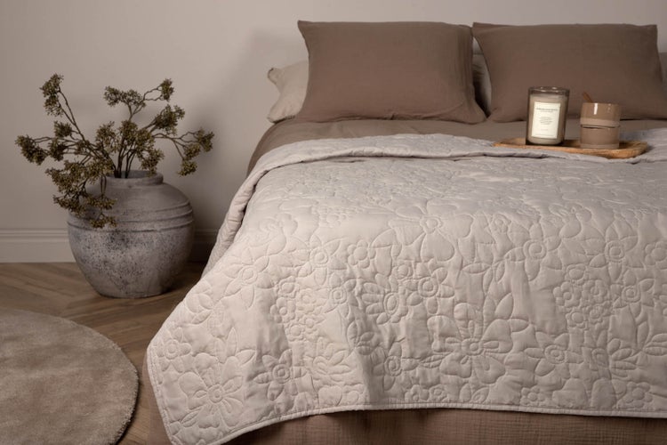 Gesteppte Tagesdecke mit Blumenmuster auf einem Bett mit Kissen, daneben eine Vase mit Zweigen.