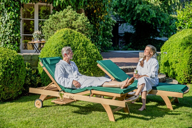 Szene mit zwei Personen in Bademänteln, die auf Gartenliegen im Garten entspannen.
