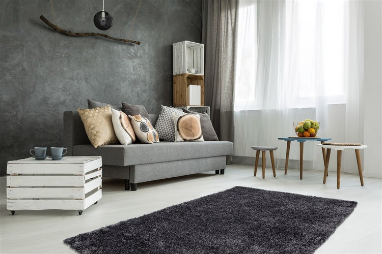 Modernes Wohnzimmer mit grauem Sofa, dunkler Strukturwand, Hochflor-Teppich, Holzkisten-Tisch und hellen Vorhängen vor großem Fenster.