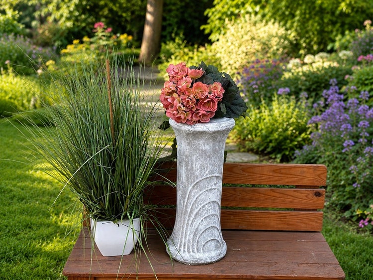 Dekorative Pflanzsäule in Steinoptik mit Blumenarrangement neben einer Kunstgras Pflanze auf einer Gartenbank.