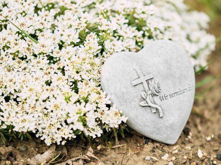 Herzförmiger Grabstein aus Stein mit eingraviertem Kreuz und Rosenmotiv sowie der Inschrift Wir vermissen dich vor blühenden Blumen