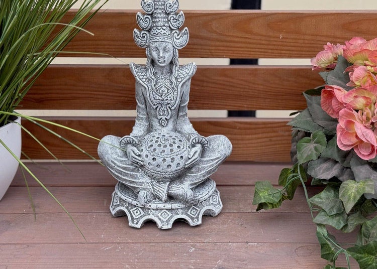 Gartenfigur einer sitzenden, meditierenden Gestalt mit kunstvollem Kopfschmuck, die eine gemusterte Kugel in den Händen hält.