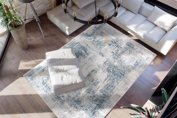 Rechteckiger Teppich mit blau-beigem Vintage-Muster in einem hellen Wohnzimmer mit weißem Ecksofa, Hocker und Parkettboden.