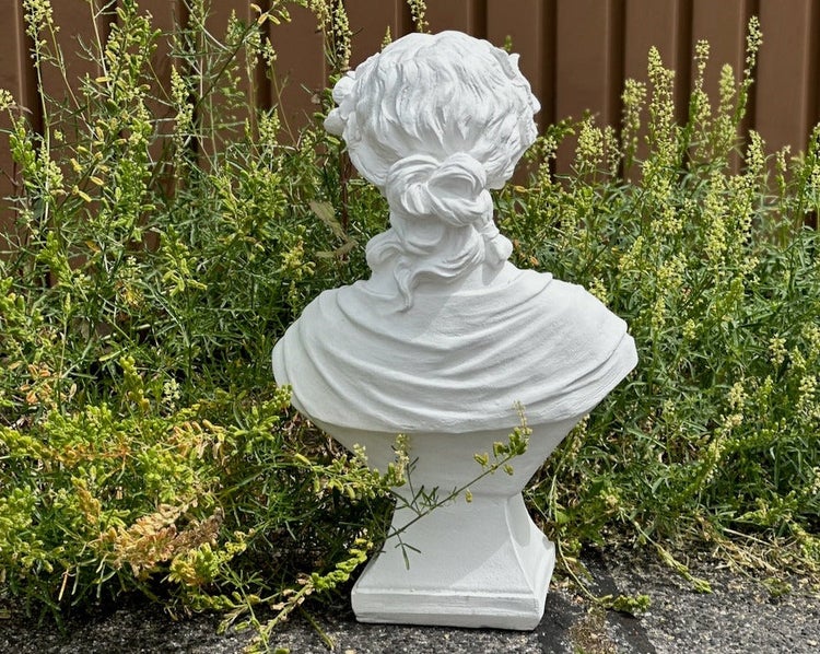 Weiße Gartenbüste einer Frau von hinten im Detail, dekorative Steinguss-Figur umgeben von grünen Pflanzen.