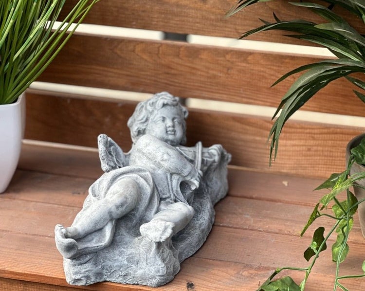 Steinfigur als liegender Engel mit Violine als Dekoration für den Garten auf einer Holzoberfläche mit Pflanzen platziert.