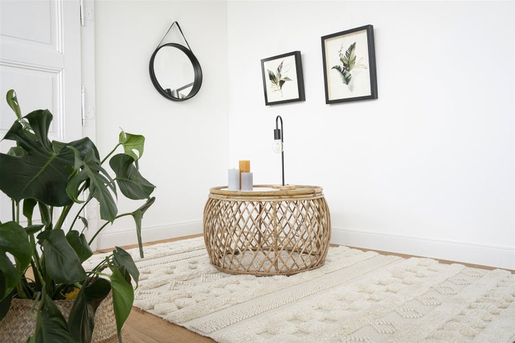 Helles Wohnzimmer mit rundem Rattancouchtisch auf einem hellen Teppich, einer Monstera-Pflanze, einem Spiegel und botanischen Wandbildern.